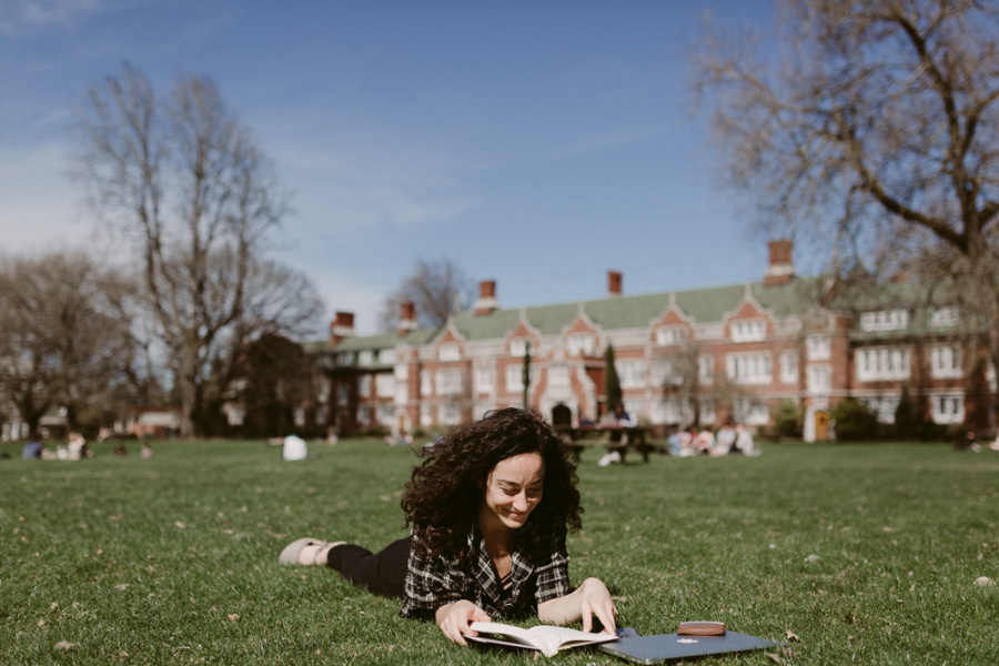 A student lays on their stomach on the grass on the Great Lawn reading a book with their laptop next to them and Old Dorm Block in the background. 