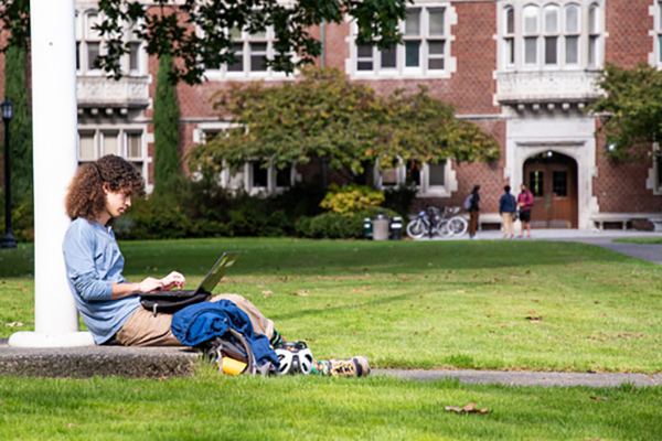 A student sits against the flag pole on the Great Lawn working on their laptop with cycling gear next to them and Eliot Hall in the background. 